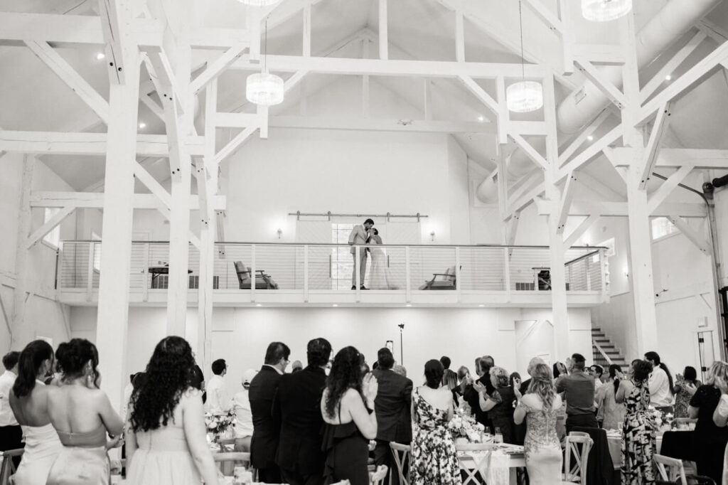 A black-and-white image of the bride and groom sharing an intimate kiss on the second-floor balcony as guests watch from below in the grand reception space.