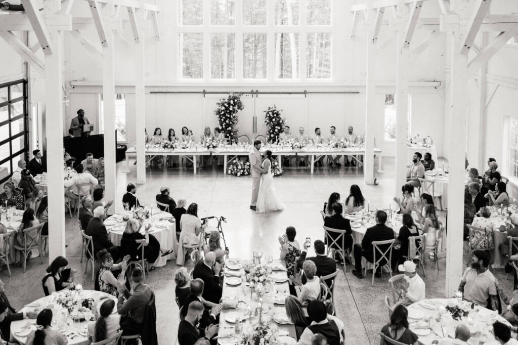 A wide-angle black-and-white view of the couple’s first dance in a bright and elegant barn, with guests seated at long reception tables and the wedding party lined up at the head table.