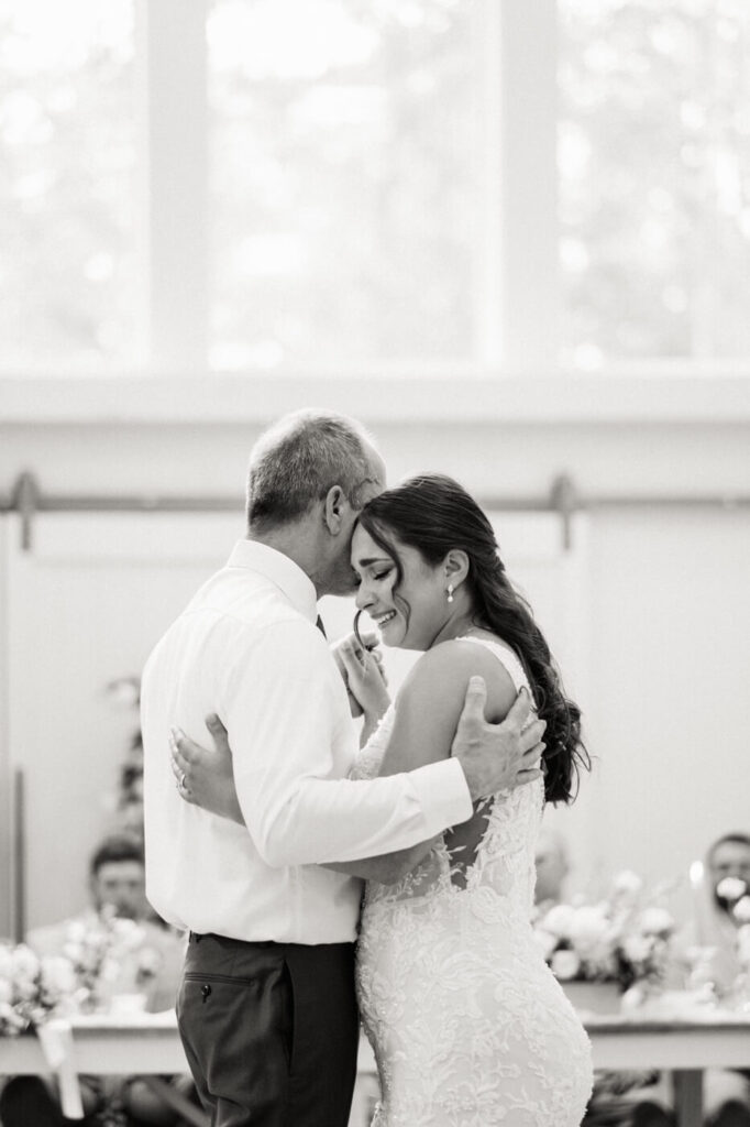 The bride shares an emotional father-daughter dance, her eyes closed as she leans into her father’s embrace, surrounded by wedding guests.