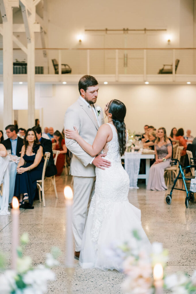 The bride and groom share their first dance, gazing at each other with candlelight and soft floral arrangements in the foreground.