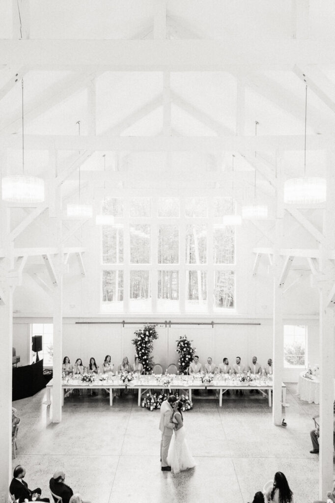 A high-key black-and-white image of the couple’s first dance in the bright and spacious reception barn, with the wedding party seated at a long head table beneath grand windows.