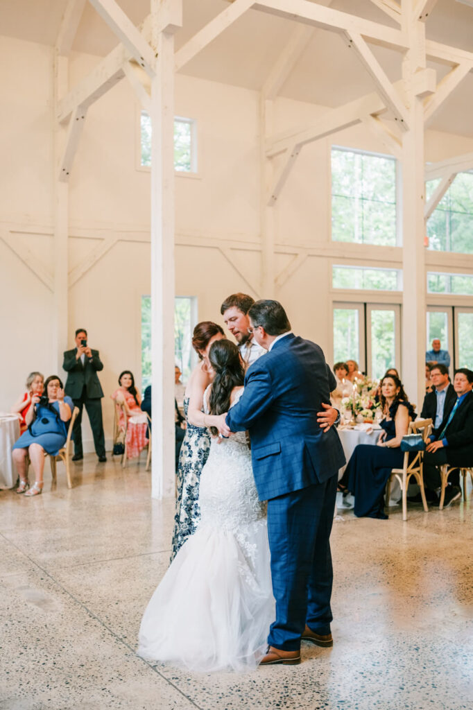 The bride and groom share a heartfelt embrace with their parents on the dance floor, surrounded by their loved ones in a bright reception space.