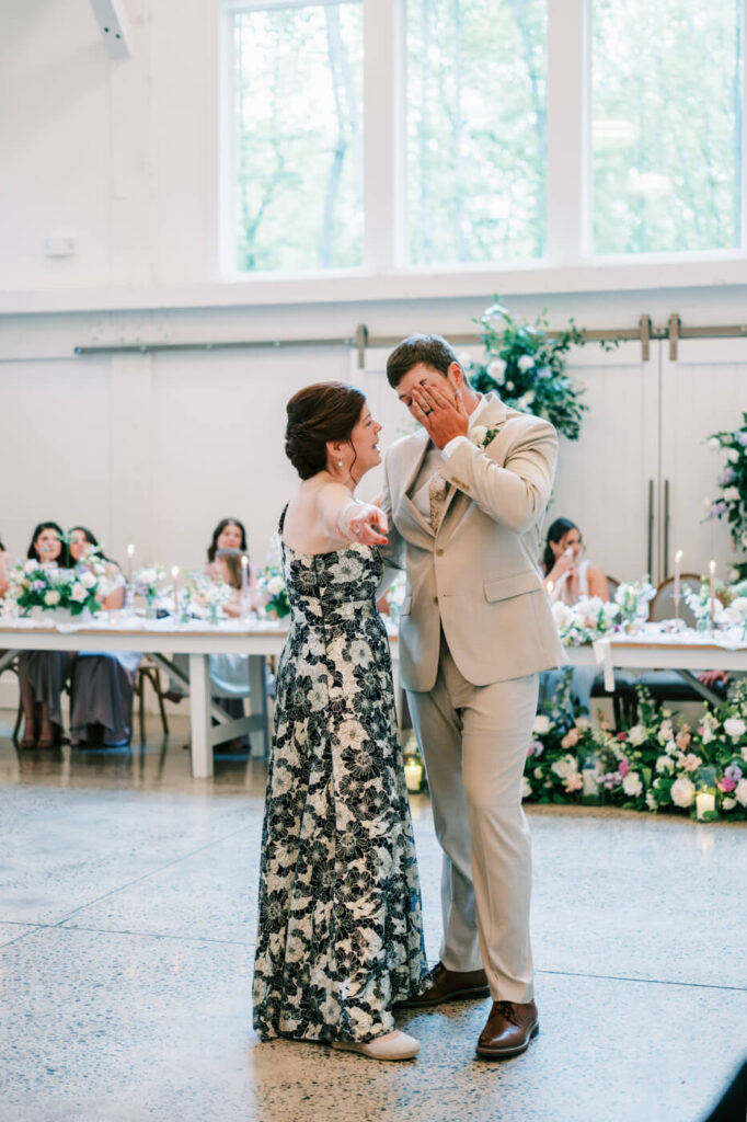 The groom wipes away tears as he shares a tender mother-son dance, his mother smiling at him as they sway together on the dance floor.