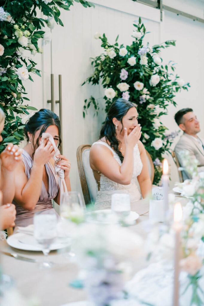 The bride and her bridesmaid wipe away happy tears while seated at the head table, floral arrangements and candlelight surrounding them.