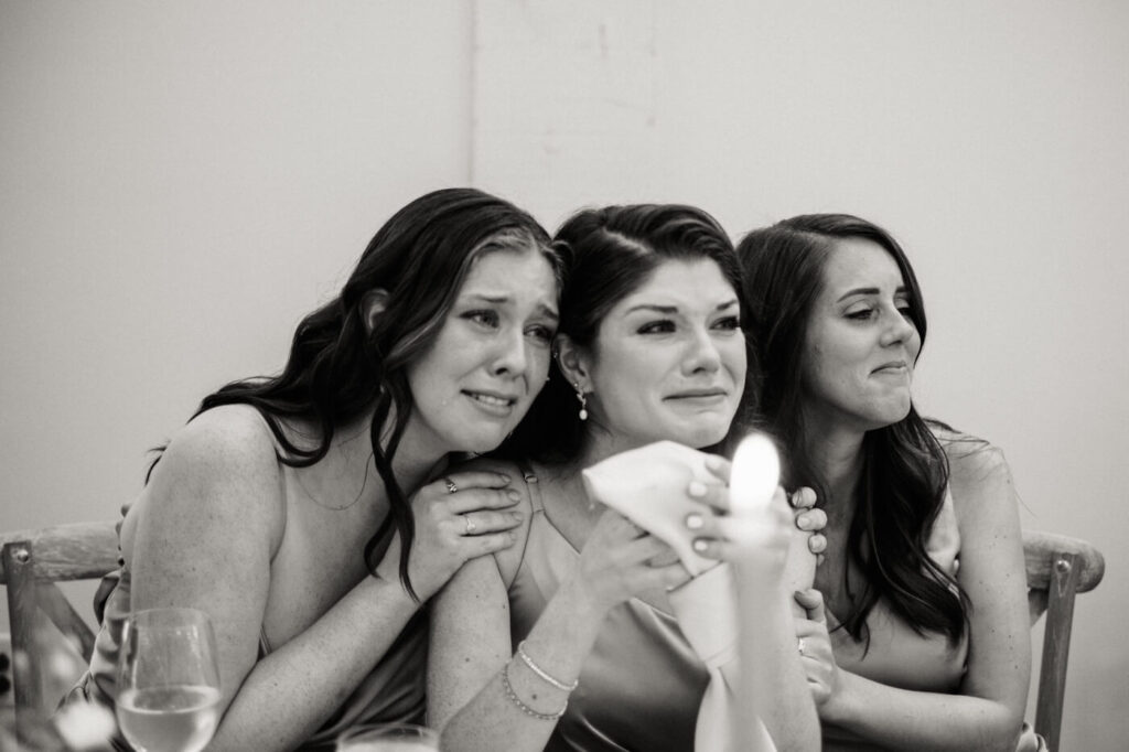 Three bridesmaids & sisters wiping tears and embracing during an emotional wedding toast at Carolina Grove.