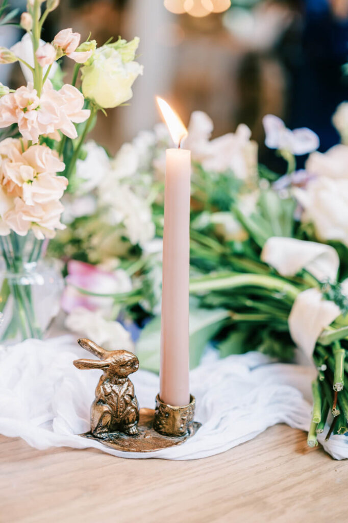 A close-up of a lit candle in a rabbit-shaped holder, surrounded by delicate florals and gauzy table decor.