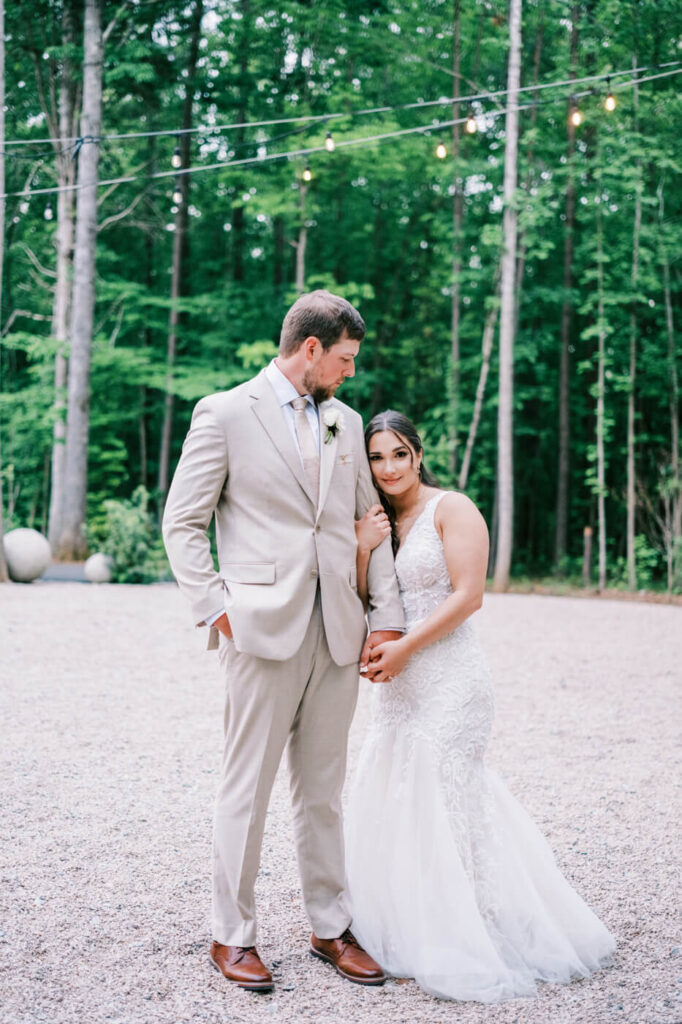 Bride and groom standing hand in hand in a peaceful wooded setting at Carolina Grove, illuminated by twinkling lights.