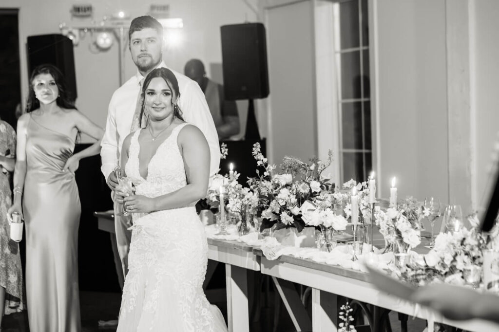 Bride and groom standing at the head table, soaking in a heartfelt speech with floral centerpieces glowing around them.