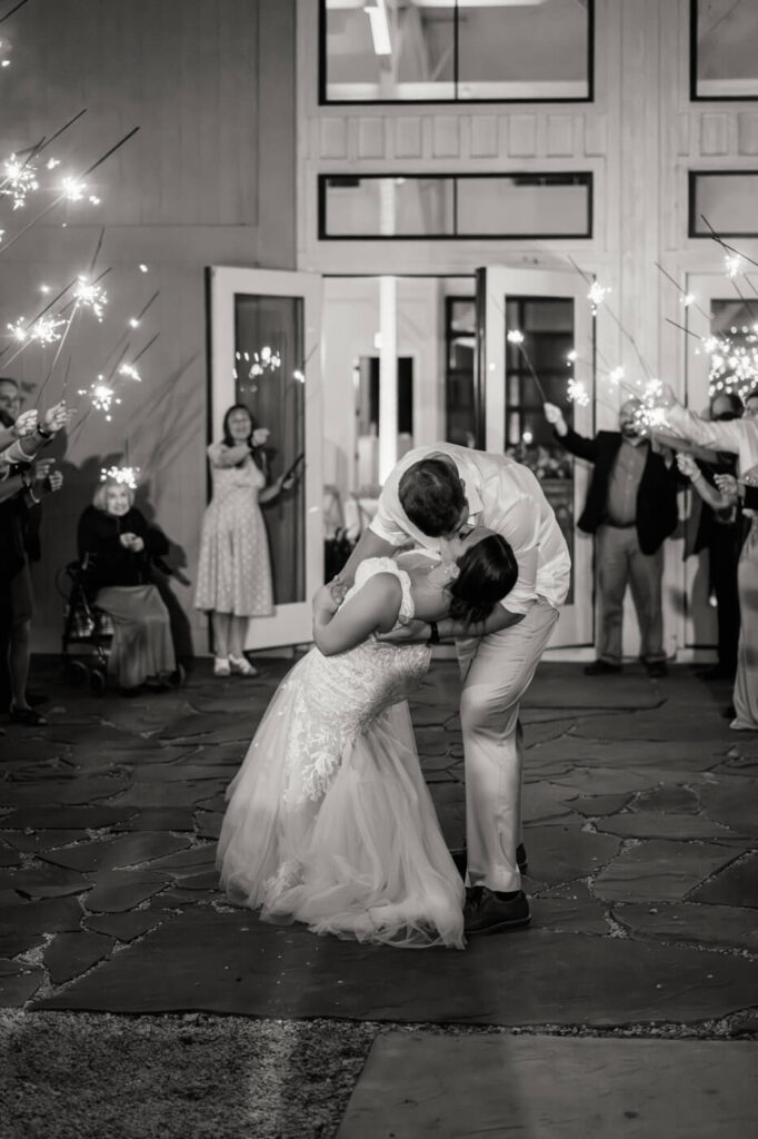 Sparkler exit with a grand kiss – The bride and groom share a passionate dip kiss while surrounded by guests holding sparklers.