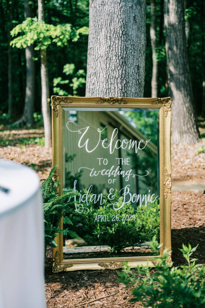 Elegant gold-framed wedding welcome sign – A beautifully designed mirror sign with calligraphy welcomes guests to Aidan & Bonnie’s wedding at Carolina Grove.