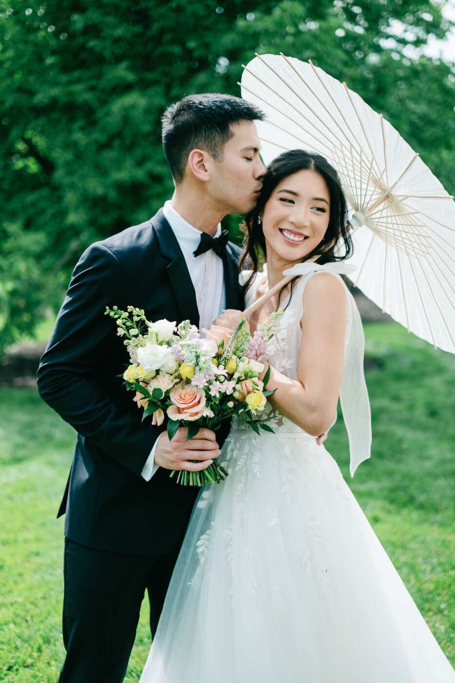 Bride and Groom with Parasol – Groom in a black tuxedo kisses bride’s forehead as she smiles, holding a bouquet of pastel flowers and a white parasol at their Sweeney Barn spring wedding.