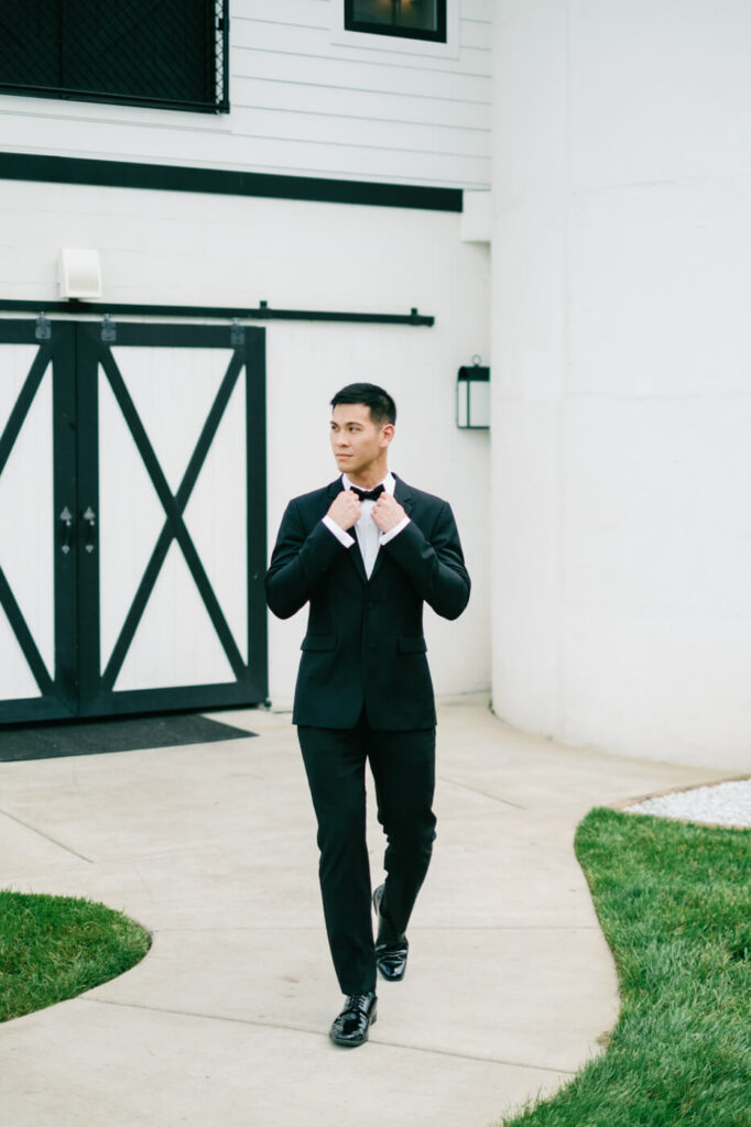 Groom Walking Outside Barn Doors – Groom adjusts his bowtie while confidently walking towards the ceremony site, framed by modern black and white barn doors at Sweeney Barn.