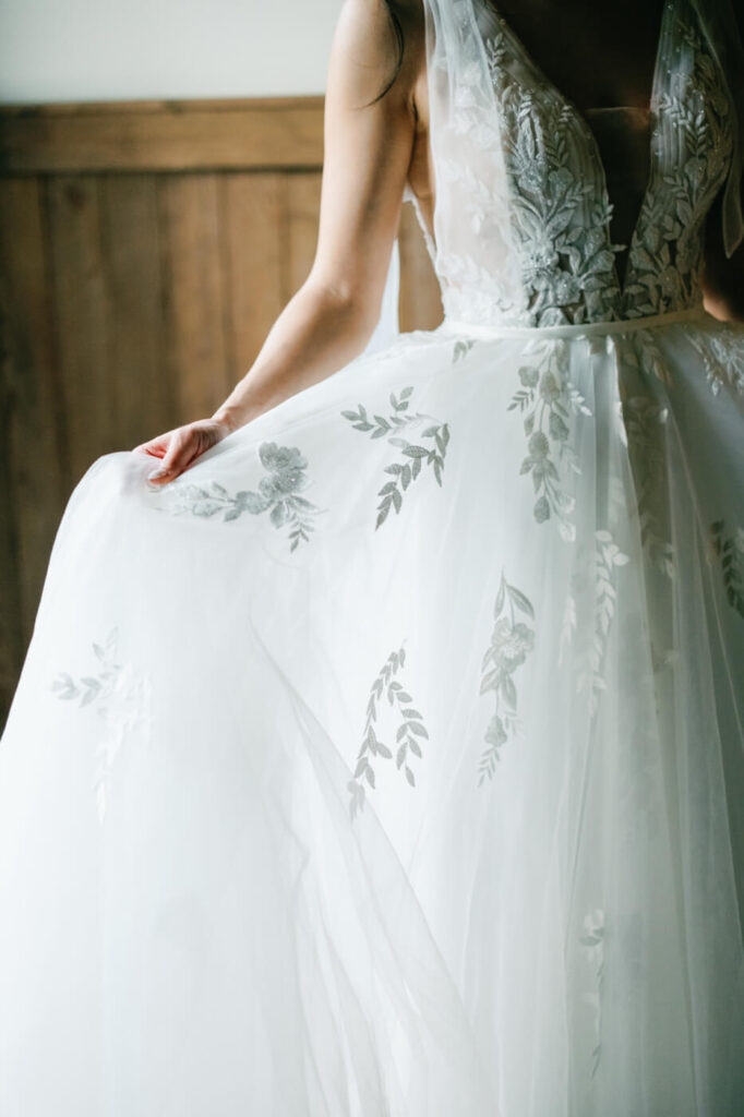 Bride Holding Wedding Gown Skirt – A romantic close-up of the bride gently lifting the lace-embroidered skirt of her wedding gown, showcasing delicate floral details.