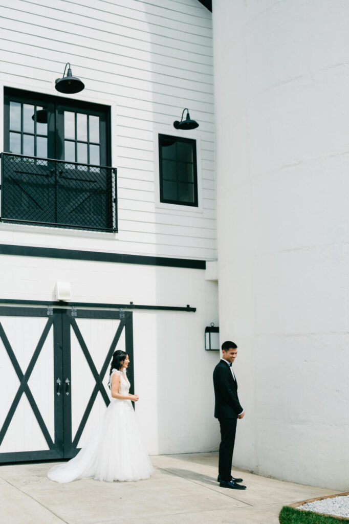 Bride Approaching Groom for First Look – The bride in a flowing white gown walking toward the groom, who stands in anticipation outside Sweeney Barn’s white barn doors.