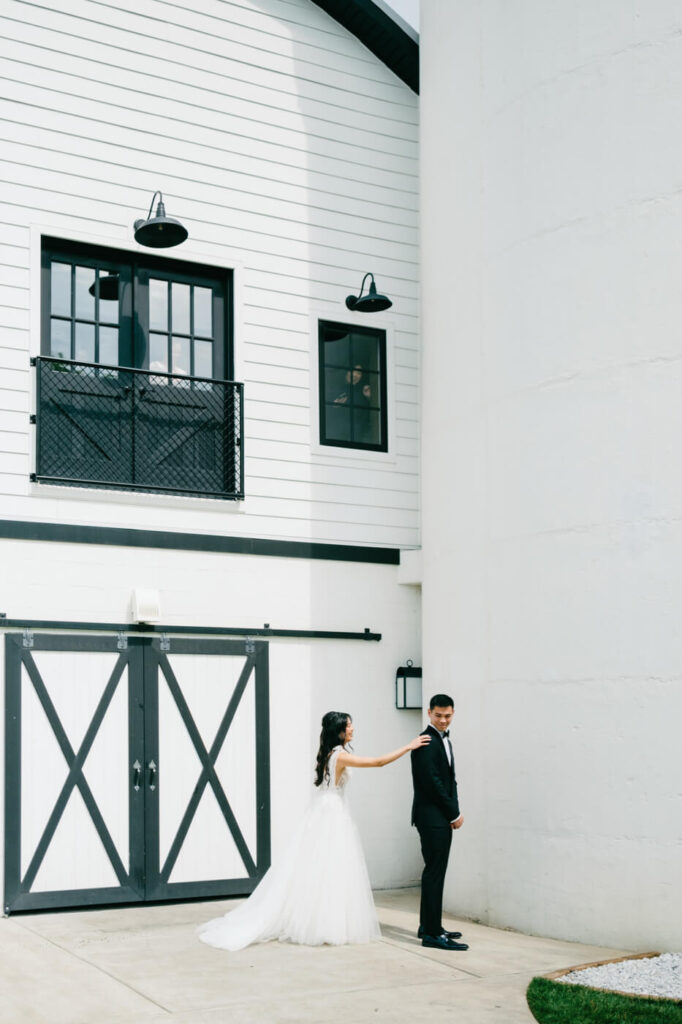 Bride Tapping Groom’s Shoulder for First Look – Emotional first-look moment as the bride reaches out to touch the groom’s shoulder, set against the elegant barn architecture.