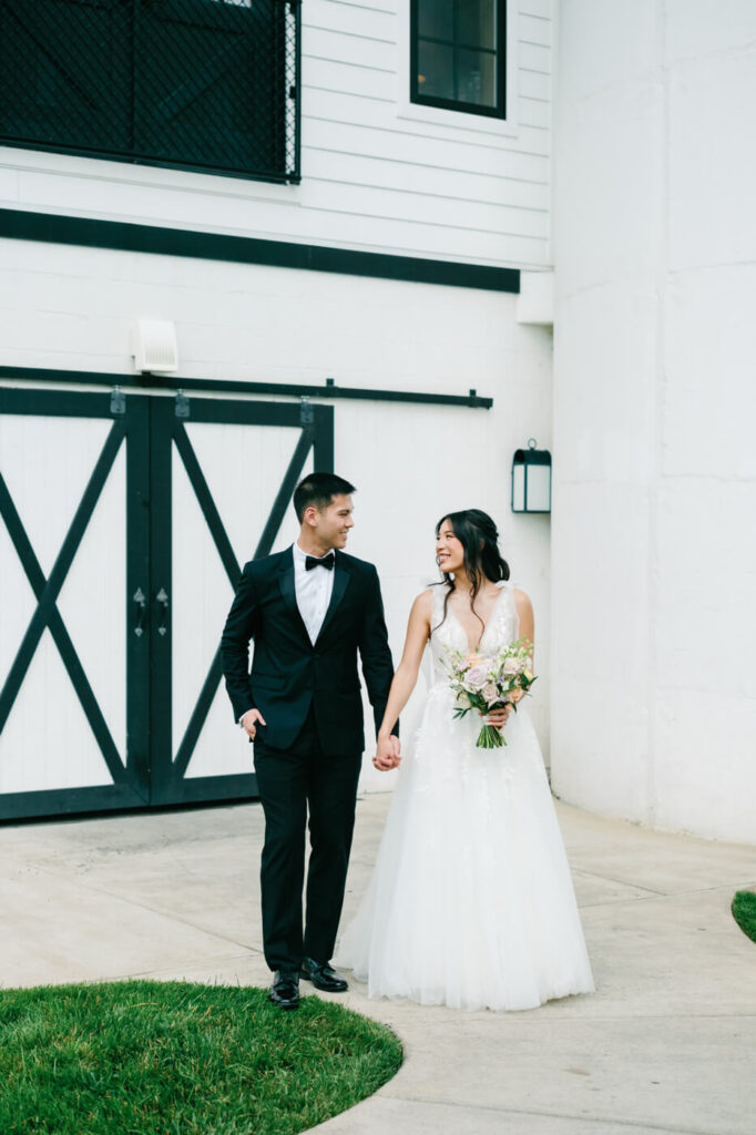 Bride and Groom Walking Hand in Hand – Newlyweds walk hand in hand outside their wedding venue, gazing at each other with love and joy.