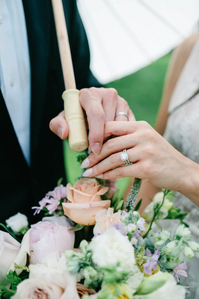 Bride and Groom Holding Hands with Wedding Rings – A close-up of the couple’s hands intertwined, showing their wedding bands and bridal bouquet in soft pastel hues.