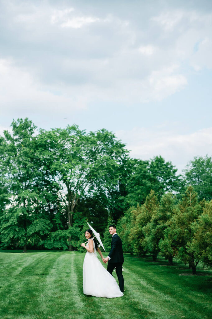 Bride and Groom Walking Through Lush Greenery – A dreamy outdoor wedding portrait of the couple strolling hand in hand through an open field, framed by vibrant green trees.