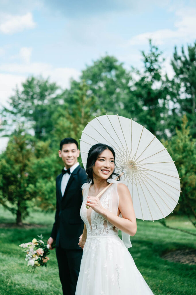 Bride with Parasol at Sweeney Barn Wedding – The bride smiles radiantly while holding a white paper parasol, with the groom following behind her, surrounded by lush greenery at Sweeney Barn.