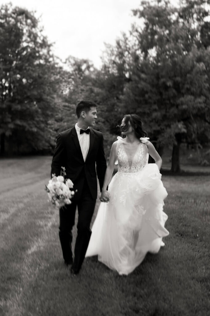 Black and White Candid Wedding Portrait – The bride and groom share a joyful moment as they walk hand-in-hand through the gardens of Sweeney Barn, with the groom holding the bridal bouquet.