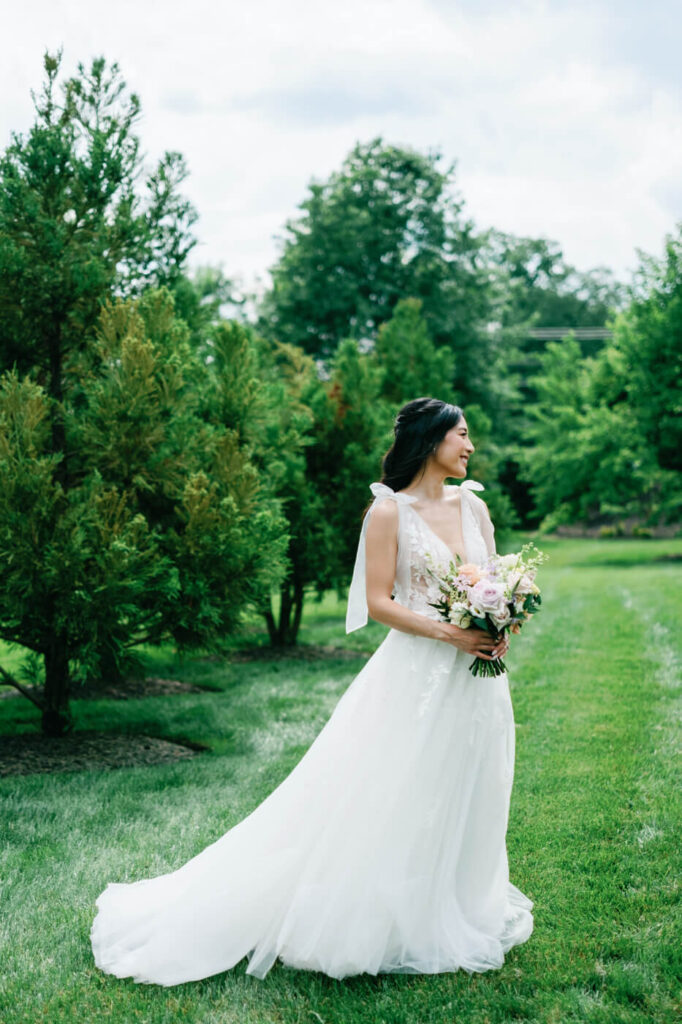 Bride Standing in Lush Greenery with Bouquet – The bride glows in her ethereal floral wedding gown, holding a pastel bouquet while standing in a picturesque garden at Sweeney Barn.