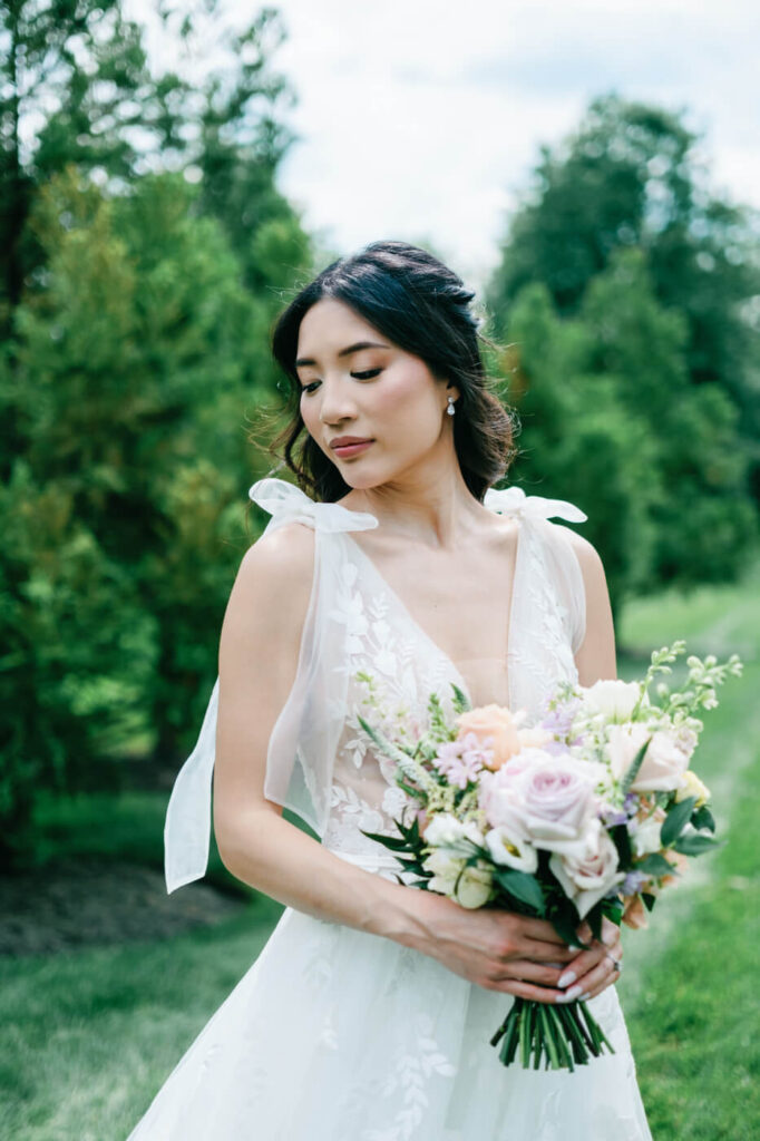 Bride Holding Floral Bouquet in Soft Natural Light – The bride looks down gracefully at her pastel floral bouquet, with her delicate lace wedding dress softly illuminated by natural light at Sweeney Barn.