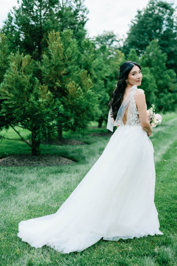 Bride Looking Over Shoulder with Elegant Train – The bride turns back with a radiant smile, showcasing her flowing wedding gown and sheer ribbon straps as she stands in a lush garden at Sweeney Barn.