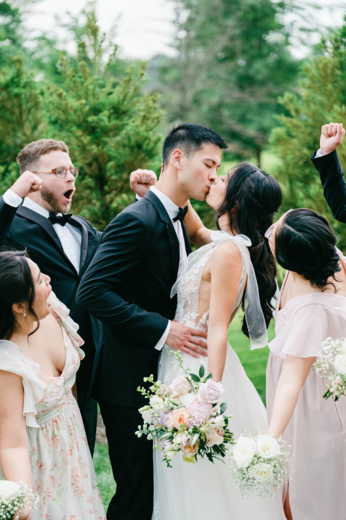 Just Married Kiss with Cheering Bridal Party – The newlyweds share a celebratory kiss surrounded by their ecstatic bridal party, set against the scenic greenery of Sweeney Barn.