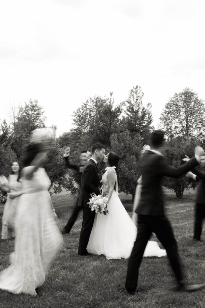 Artistic Motion Wedding Portrait with Guests – A timeless black-and-white image of the bride and groom kissing while their wedding party joyfully moves around them, captured in motion blur.