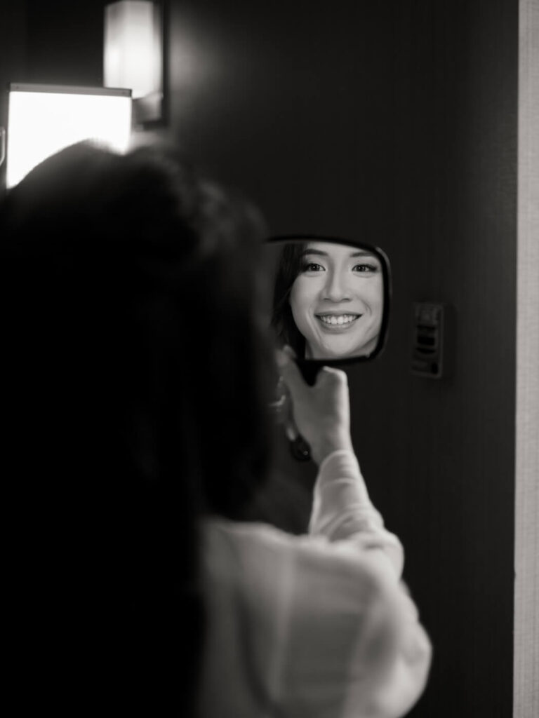 Bride Looking in Mirror – Candid black and white image of the bride smiling at her reflection while holding a small mirror, a quiet moment before her Sweeney Barn wedding ceremony.