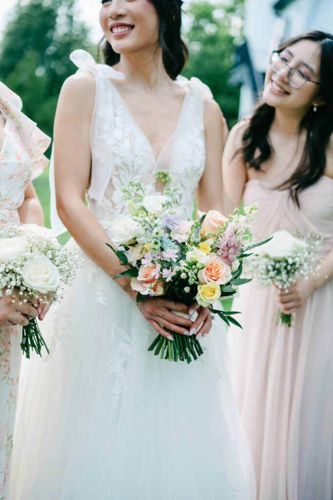 Bride holding her bouquet with bridesmaids nearby – A joyful bride in a lace wedding gown holds a pastel floral bouquet while her bridesmaids in soft pink dresses stand beside her, smiling.