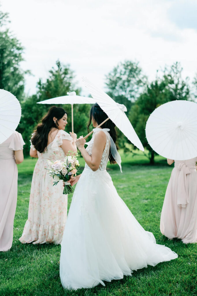 Bride and bridesmaids walking with parasols – The bride and her bridesmaids walk through a lush green field, carrying elegant white paper parasols and holding their bouquets.