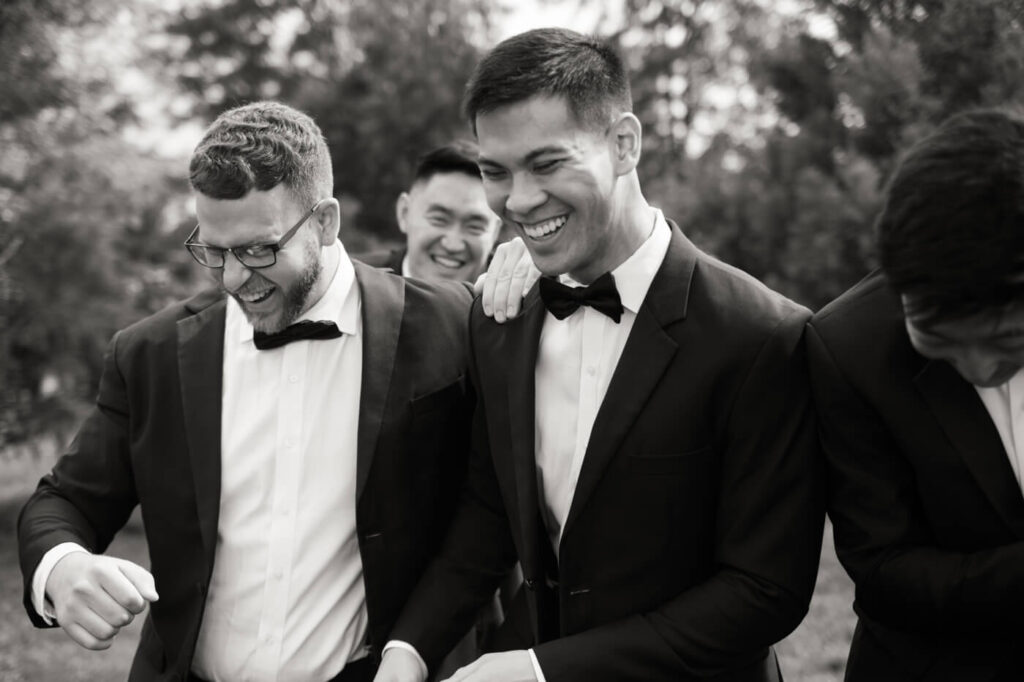 Groom laughing with groomsmen – The groom and his groomsmen, dressed in black tuxedos, share a candid moment of laughter in an outdoor setting.