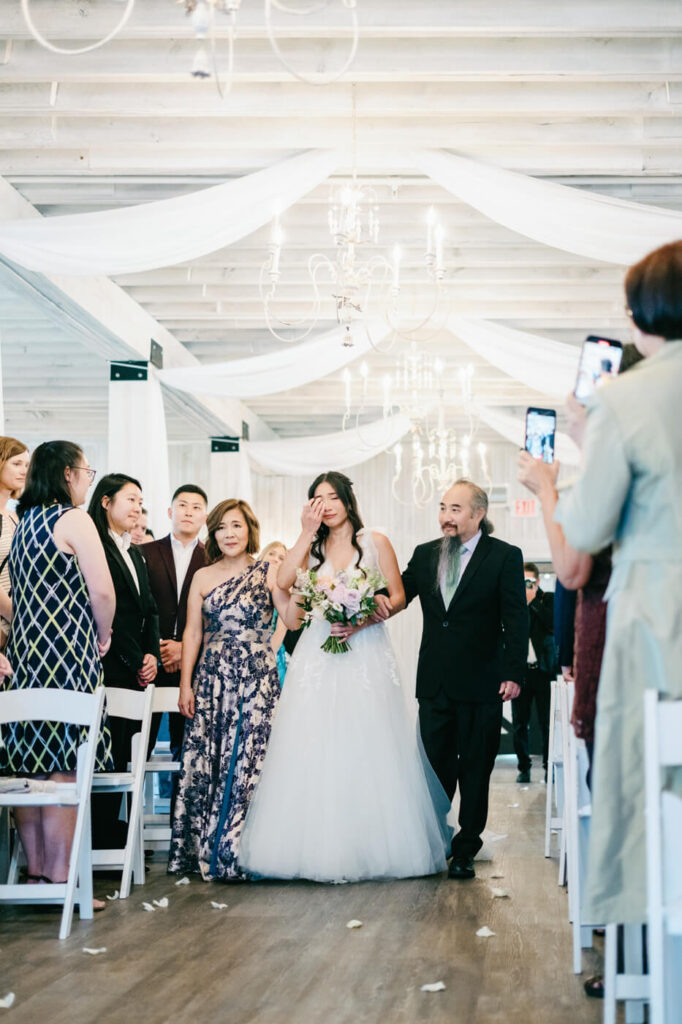 Bride walking down the aisle with her parents – An emotional bride wipes away a tear as she walks arm-in-arm with her parents down a beautifully draped aisle, surrounded by guests.