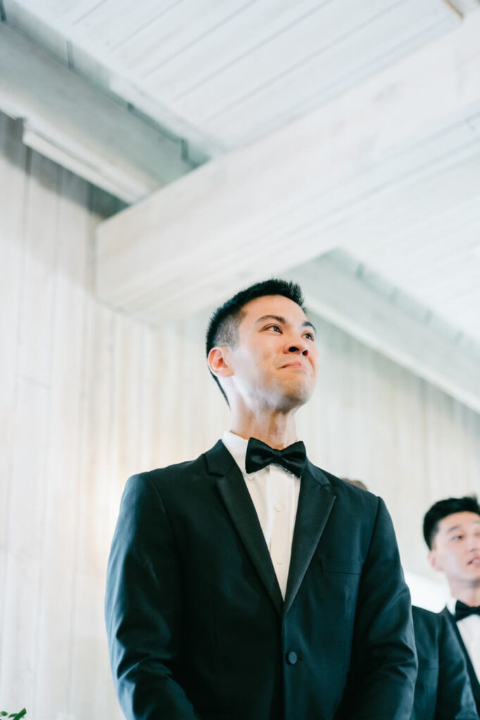 Groom’s emotional reaction during the ceremony – The groom, dressed in a black tuxedo, fights back tears as he watches his bride walk down the aisle.