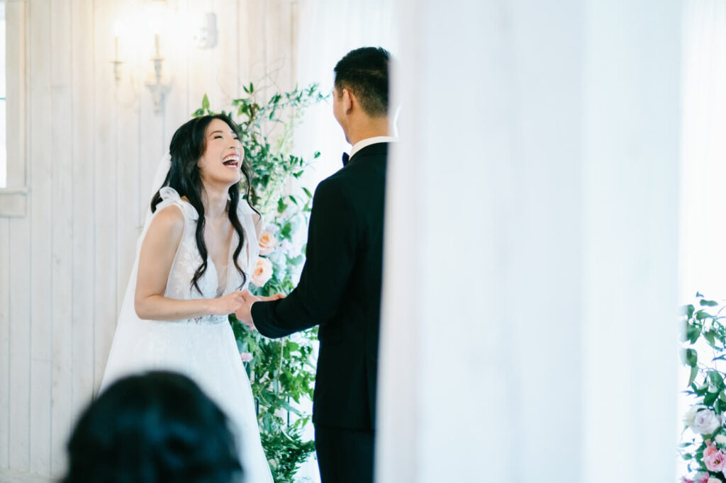 Bride and groom laughing during their vows – A heartfelt moment as the bride and groom share laughter during their wedding vows, surrounded by floral arrangements and soft white drapery.