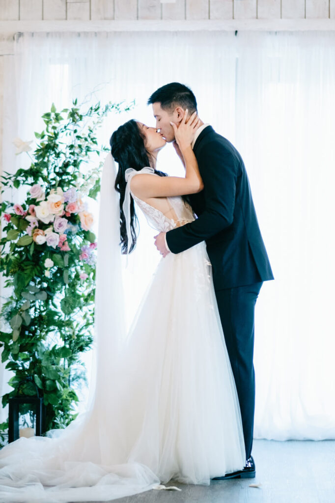 Bride and groom’s first kiss – The newlyweds share a romantic first kiss in front of a floral-adorned ceremony backdrop, bathed in soft natural light.