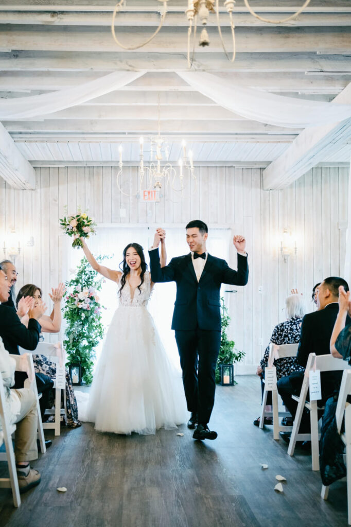 Bride and groom celebrating their marriage – The bride raises her bouquet in triumph while holding her husband’s hand as they walk down the aisle together, surrounded by cheering guests.