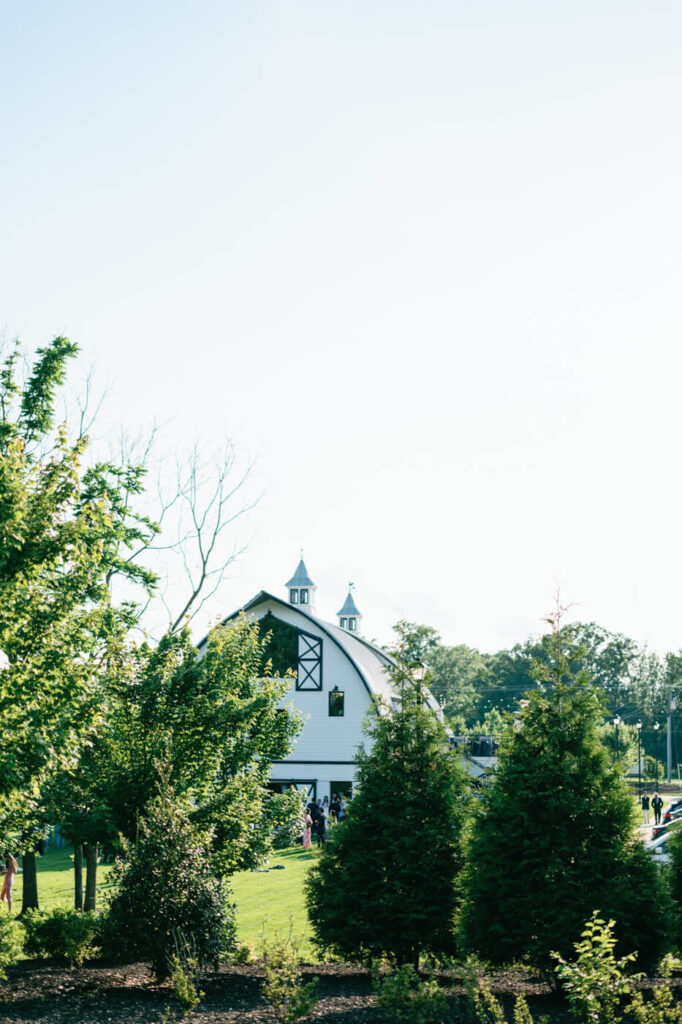 Exterior of Sweeney Barn wedding venue – A charming white barn with black accents stands surrounded by lush greenery under a clear blue sky, setting a picturesque scene.