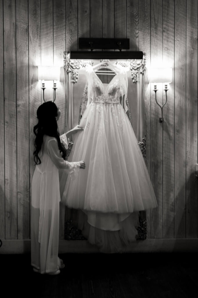 Bride with Wedding Dress – Bride in a sheer bridal robe gently touching her lace wedding gown hanging in an ornate mirror, preparing for her spring wedding at Sweeney Barn.