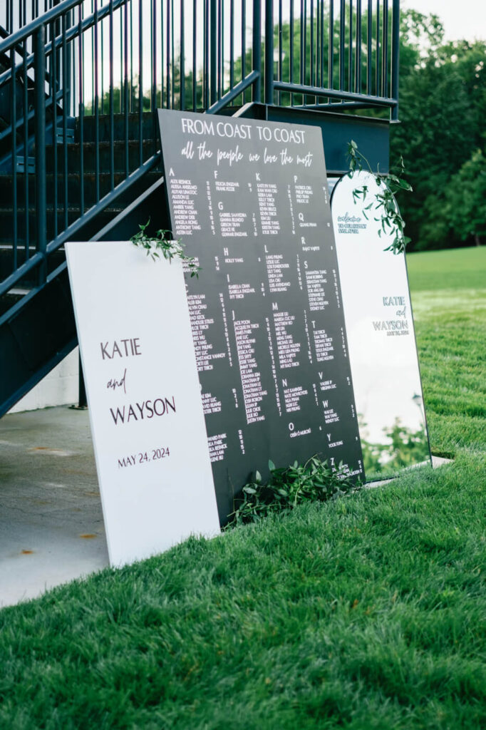 Wedding seating chart with guest names – A modern black and white wedding seating chart displays guests' names, adorned with greenery and elegant calligraphy, placed against a staircase.