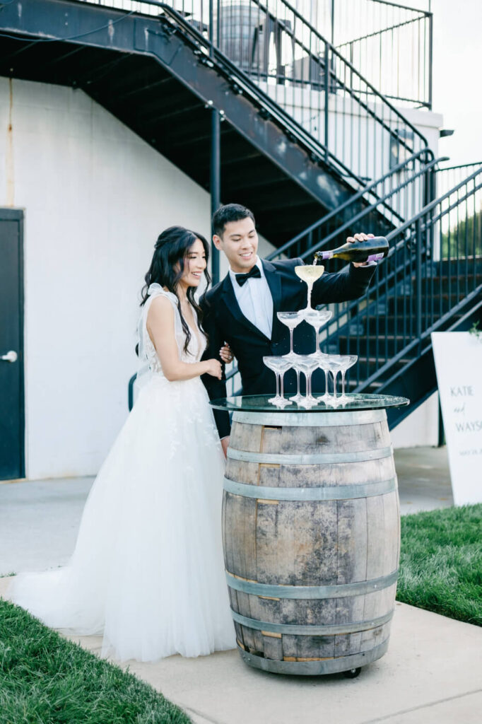 Bride and groom pouring champagne tower – A newlywed couple stands together, smiling as the groom pours champagne into a cascading tower of coupe glasses set on a rustic barrel.