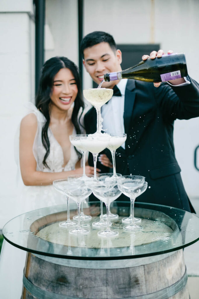 Close-up of champagne pouring – A joyful bride and groom watch as sparkling champagne overflows from the top glass of a tiered champagne tower, creating an elegant celebratory moment.