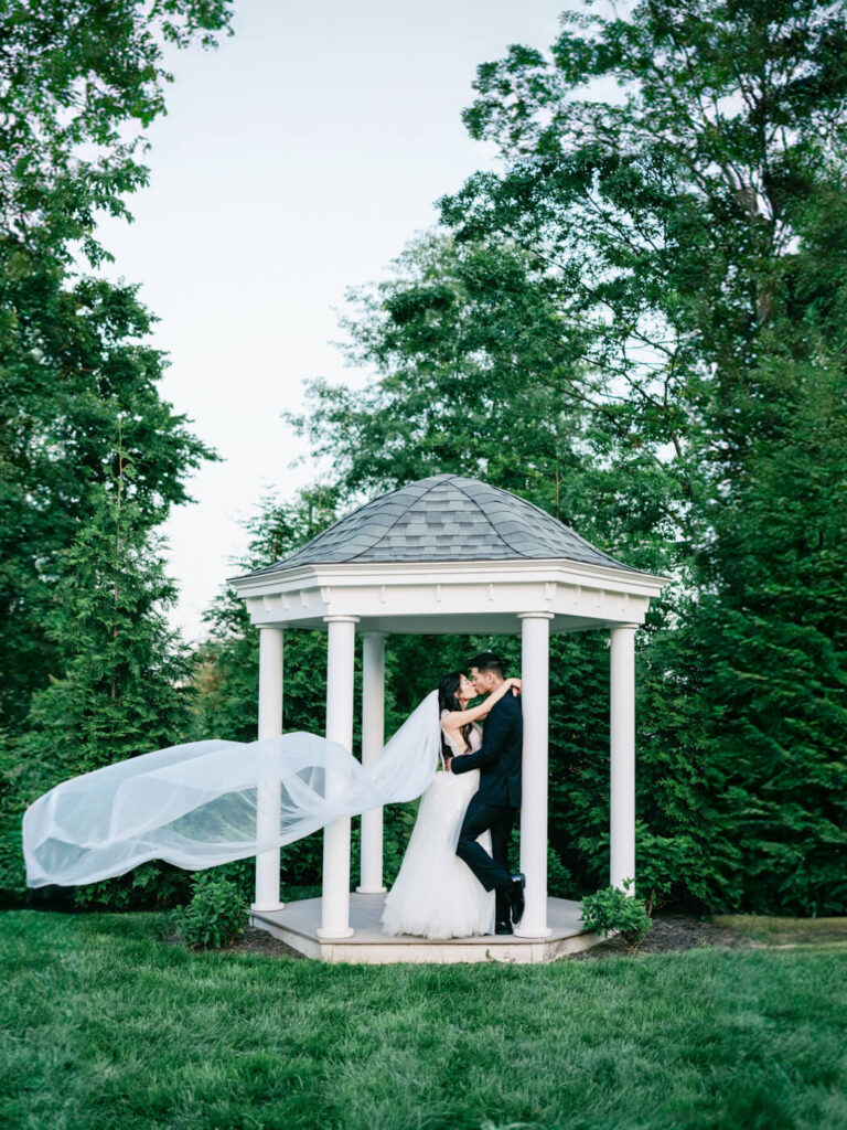 Romantic gazebo embrace – The bride and groom share a passionate kiss under a charming white gazebo, with the bride’s veil flowing in the breeze against a lush green backdrop.