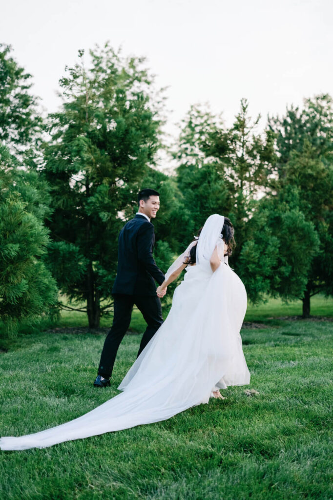 Bride and groom walking hand in hand – The newlyweds stroll through a picturesque garden, holding hands as the bride lifts her gown slightly, her long veil trailing behind her.