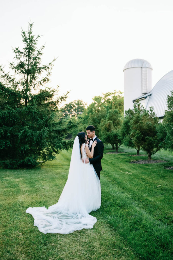 Bride and groom in an intimate embrace near a barn – The couple stands in a sunlit garden, gazing into each other’s eyes with the iconic curved barn silo in the background.