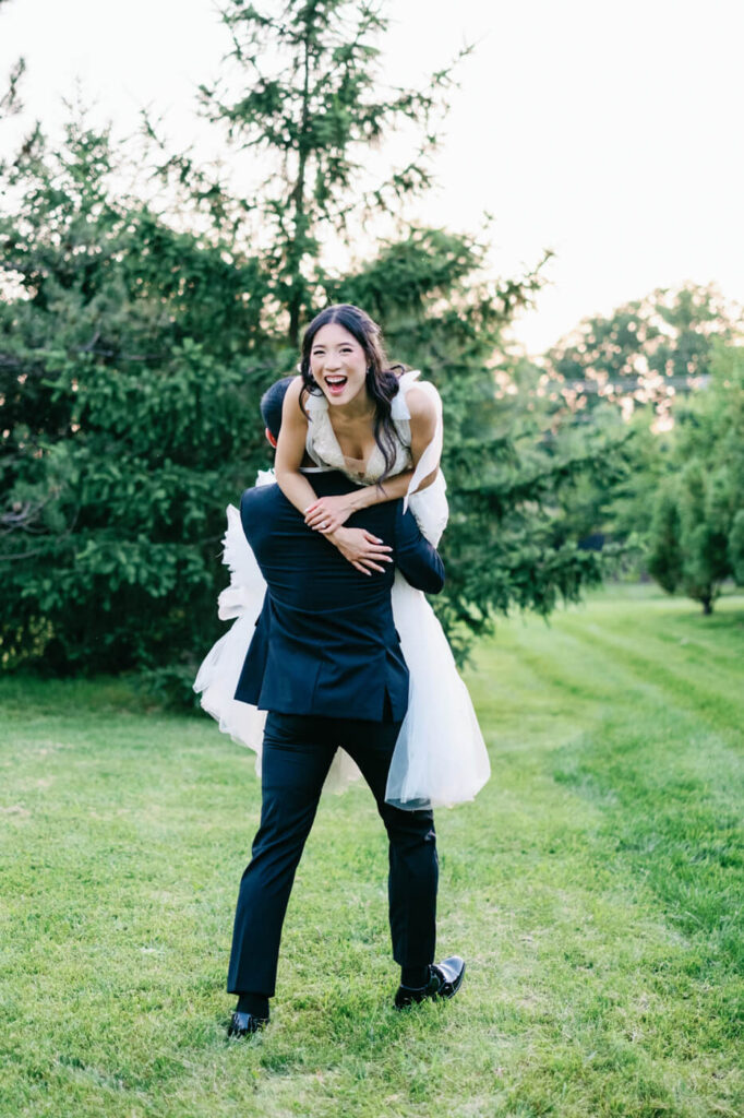 Groom playfully lifting the bride – The groom picks up the bride in a joyful embrace, while she laughs with delight in a green field surrounded by trees.