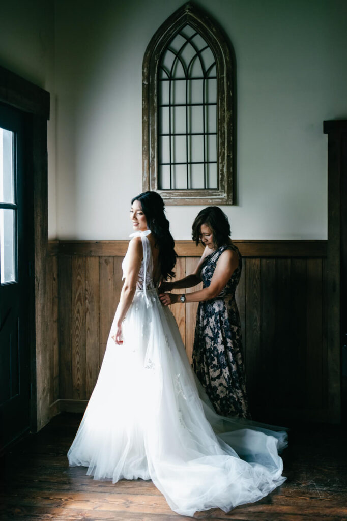 Mother Helping Bride into Dress – Bride in a delicate lace gown smiles as her mother fastens the back, captured in soft natural light at Sweeney Barn.