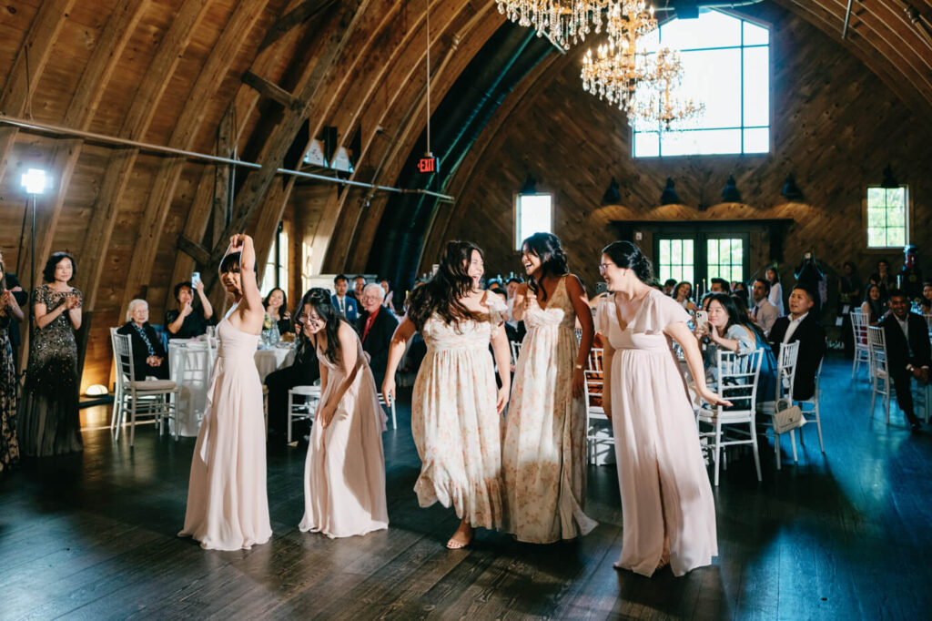 A joyful moment as a group of bridesmaids in blush and floral dresses laugh together while walking across the reception floor at Sweeney Barn, surrounded by guests in the warmly lit barn venue.