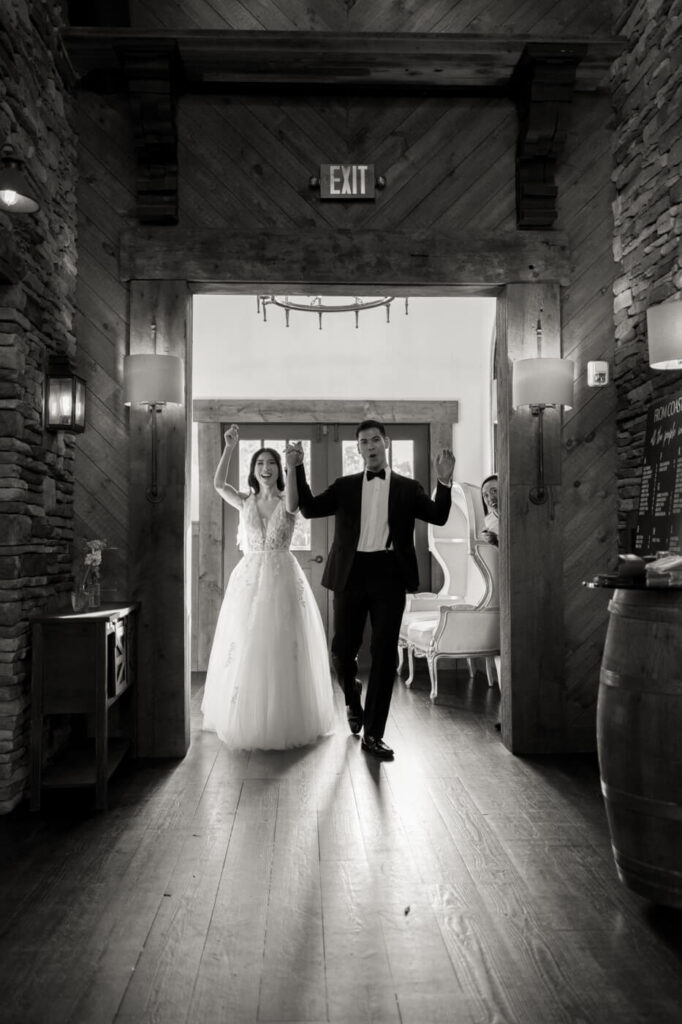 A black-and-white photograph of the newlyweds making a grand entrance into their reception, hand in hand, with bright smiles, framed by rustic wooden beams and stone walls.